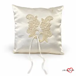 Off-white satin ring cushion with floral lace appliqués, ribbon ties and a small brown mark near the upper edge